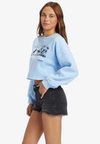Felpa cropped di colore azzurro con maniche lunghe e stampa grafica di palme e uccelli sul davanti, abbinata a pantaloni corti in denim neri.