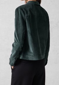 Dark green corduroy jacket with a collar, visible seam details, and side pockets, styled with black trousers. Soft textured fabric.