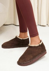 Pantofole in pelle scamosciata marrone con bordatura in pelliccia crema e cuciture decorative lungo la suola. Design a taglio basso adatto per l'uso interno.