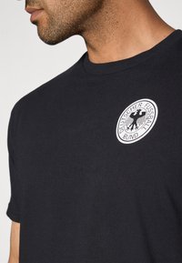 Mustast puuvillasest T-särgist, millel on vasakul rinnal valge ringikujuline Saksa Jalgpalliliidu logo tikand.