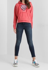 Koraal kleurige cropped hoodie met een tijgerafbeelding, blauwe skinny jeans met zijstrepen en chunky sneakers met multikleurige accenten.