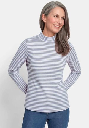 Femme aux cheveux gris souriante, portant un col roulé à manches longues rayé violet clair et blanc et un jean bleu, debout les mains derrière le dos.