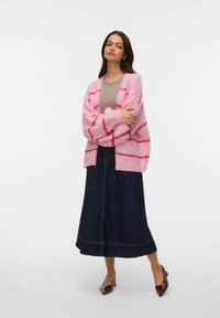 Ροζ πλεκτή ζακέτα με κόκκινες ρίγες, φορεμένη πάνω από μπεζ τοπ, συνδυασμένη με μακριά σκούρα τζιν φούστα και καφέ παπούτσια με μυτερό μύτο.