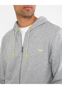 Sudadera con cremallera gris y capucha ajustable. Presenta detalles amarillos en los cordones y un pequeño logo verde en el pecho. Textura suave.