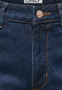 Artėjimas prie tamsiai mėlynų džinsų priekinės dalies su vario spalvos sagomis ir oranžinėmis siūlėmis.