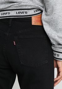 Los jeans de mezclilla negros tienen un bolsillo trasero con la etiqueta del logo de Levi's, combinados con una camisa de manga larga gris con una cinturilla de Levi's en blanco y negro.