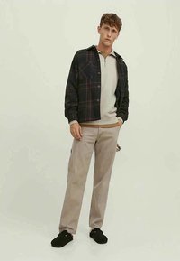 Schwarze Karoshemdjacke über einem hellgrauen Zip-Up-Oberteil, kombiniert mit cremefarbener Hose und schwarzem Schuhwerk. Einfaches Design und neutrale Farben.