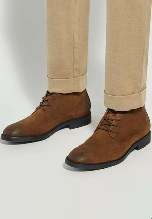 Brown leather ankle boots with a round toe, laced front, and smooth texture, paired with beige tapered trousers, featuring rolled cuffs.
