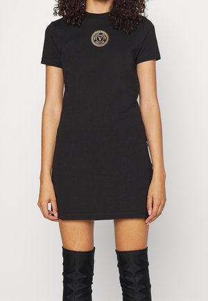 Femme portant une mini-robe noire à manches courtes avec un logo rond doré sur la poitrine et des bottes noires jusqu'aux genoux, se tenant contre un fond blanc.