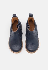 Námornícky kožené boty s hladkou texturou, detailem brogue, bočním zipem a světlými hnědými podrážkami. Obsahují dva úchyty pro snadné obutí.