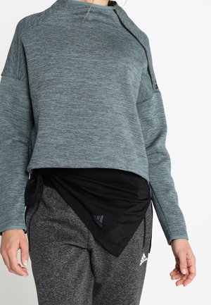 Kobieta ubrana w szarą sportową bluzkę z długim rękawem, czarny szalik i szare spodnie dresowe Adidas z białym logo, stoi na jednolitym białym tle.