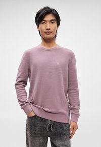 Lilla sweater med et struktureret mønster, rund hals og lange ærmer, parret med mørke jeans. Diskret logo detalje på brystet.