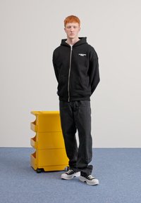 Črna zip-up kapuca z belim napisom, temne kavbojke in črno-bele teniske, stoji ob rumeni skladiščni enoti na modri podlagi.