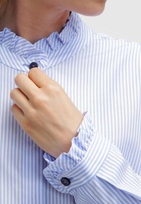 Niebieska i biała bluzka w paski z falbankowym kołnierzykiem, zapięciem na guziki przy mankietach oraz gładkim materiałem. Posiada mały, ciemny guzik przy kołnierzyku.