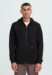 Haut à capuche zippé noir en mélange de coton, doté d'une poche avant, d'une capuche avec cordon de serrage et d'un tissu texturé. Porté par-dessus un t-shirt blanc.