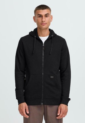 Haut à capuche zippé noir en mélange de coton, doté d'une poche avant, d'une capuche avec cordon de serrage et d'un tissu texturé. Porté par-dessus un t-shirt blanc.