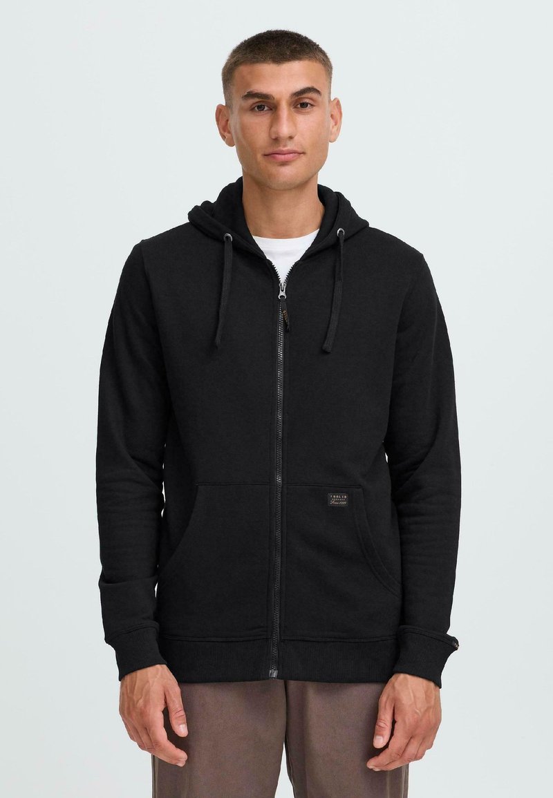 Haut à capuche zippé noir en mélange de coton, doté d'une poche avant, d'une capuche avec cordon de serrage et d'un tissu texturé. Porté par-dessus un t-shirt blanc.