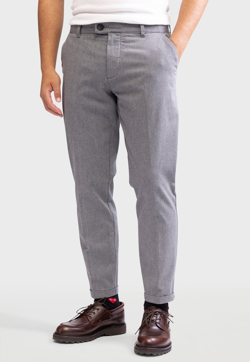 Homme portant un pantalon gris sur mesure, des chaussettes noires avec un motif de cœur rouge, et des chaussures marron foncé à lacets, debout avec une main dans la poche.
