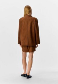 Veste en suède marron à coupe ajustée, dotée d'une couture arrière unique, portée par-dessus une mini-jupe marron assortie et des chaussures plates noires.