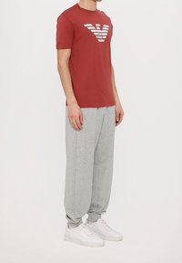 Czerwona bawełniana koszulka z białym paskowanym logo, zestawiona z szarymi dresami. Model nosi białe sneakersy, strój o luźnym kroju.