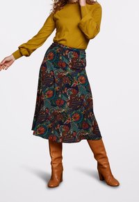 Jupe midi à motif floral sur fond sombre, arborant des couleurs vives, associée à un haut à manches longues moutarde et des bottines à talons marron.
