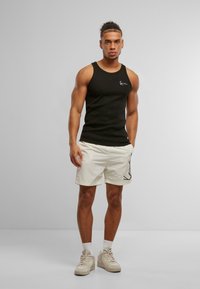 Débardeur noir côtelé avec logo blanc ; shorts clairs avec un motif abstrait noir ; baskets beiges et chaussettes blanches.