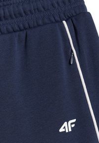 Pantalon de sport bleu marine avec taille élastique, bande blanche latérale, poche zippée et logo blanc « 4F » près de l'ourlet.