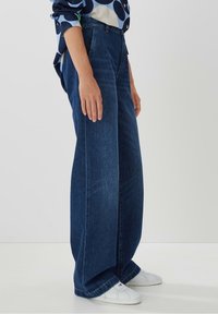 Breda mörkblå denimjeans med hög midja, utrustade med sidofickor och en ren fåll. Matchas med vita sneakers.