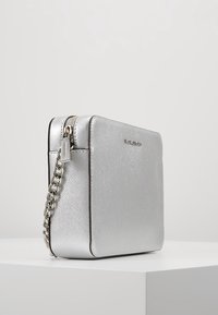 Bolso rectangular de plata texturizado con una correa de cadena metálica y cierre de cremallera, colocado sobre una superficie blanca con un fondo neutro.