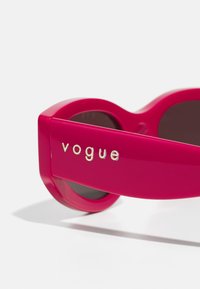 Ochelari de soare roz aprins cu un design rotund, având lentile întunecate și un logo argintiu pe braț, material plastic neted și o formă robustă.