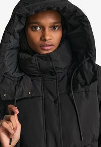 Svart vadderad jacka med hög krage och stor huva. Har en glänsande yta, tryckknappar och justerbara dragsnören för en säker passform.