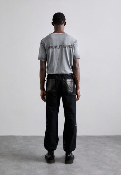 Graues Baumwoll-T-Shirt mit "HELMut LANG"-Logo auf dem Rücken, kombiniert mit schwarzen Hosen mit Lederentaschen und Reißverschlüssen. Schwarze Schuhe.