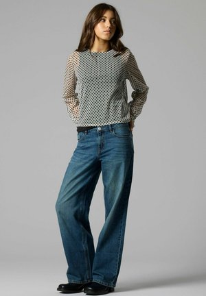 Jeune femme portant un haut transparent à manches longues à pois, un jean bleu à jambes larges et des chaussures noires, debout avec les mains derrière le dos.