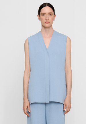 Vrouw met donker haar en een middenscheiding draagt een lichtblauw mouwloos gilet en bijpassende wijde broek tegen een effen lichte achtergrond.