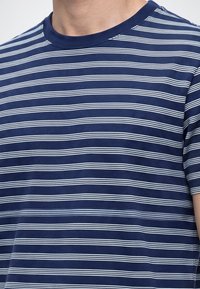 Personne portant un t-shirt bleu marine avec de fines rayures horizontales blanches et un col rond, montrant le haut du torse et une partie d'un bras.