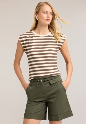 Joven mujer con largo cabello rubio que lleva una camiseta sin mangas a rayas beige y marrón y pantalones cortos verdes oliva con cinturón y las manos en los bolsillos.