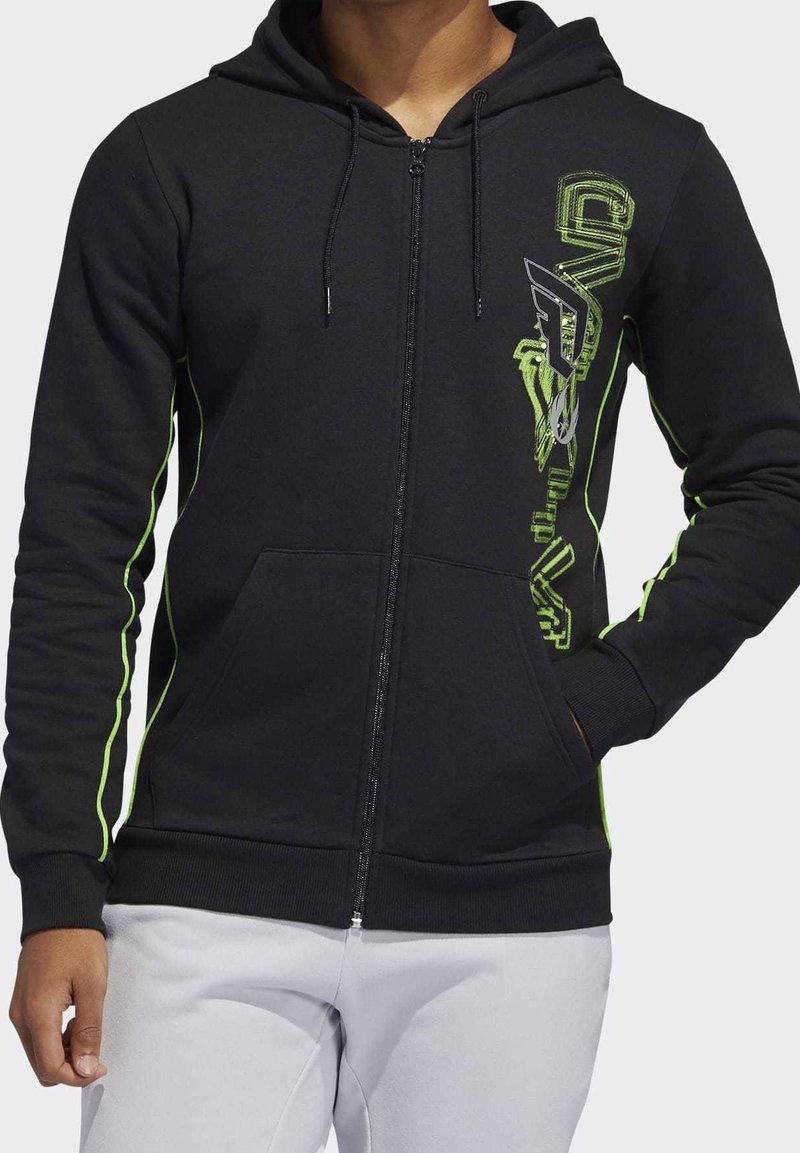 Personne portant un sweat à capuche noir zippé avec des coutures vertes et un texte graphique sur le côté droit, associé à un pantalon de couleur claire.