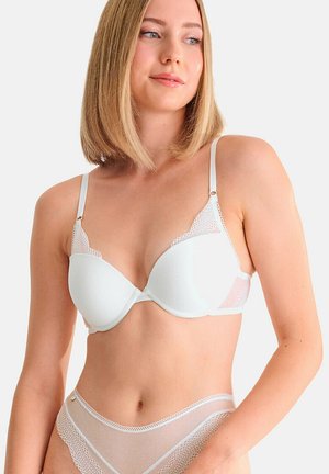 Femme aux cheveux blonds mi-longs portant un soutien-gorge blanc orné de dentelle et une culotte taille haute en filet assortie, sur un fond uni.