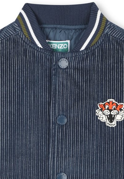 Veste en velours côtelé bleu avec col rayé côtelé, boutons-pression et patch brodé d'un visage de tigre orange souriant sur la poitrine.