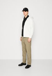 Felpa bianca con zip, maglia nera sotto, pantaloni beige, scarpe nere e un cappellino nero con logo. Design semplice e moderno con colori neutri.