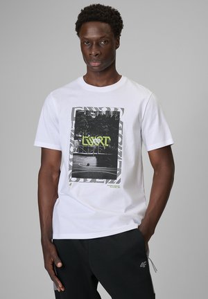 Młody mężczyzna o ciemnej karnacji i krótkich kręconych włosach, ubrany w biały t-shirt z nadrukiem i czarne spodnie na szarym tle.