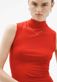 Top sin mangas de cuello alto en rojo, confeccionado en una tela suave y de ajuste ceñido. Acompañado de un sencillo collar de oro con un colgante en forma de letra.