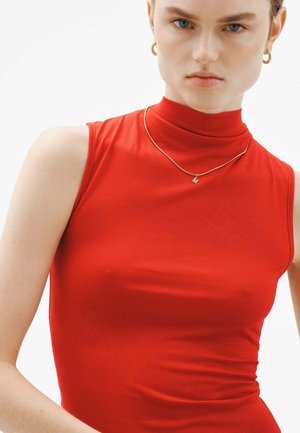 Top sin mangas de cuello alto en rojo, confeccionado en una tela suave y de ajuste ceñido. Acompañado de un sencillo collar de oro con un colgante en forma de letra.