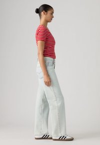 Κοντομάνικο τοπ με κόκκινες και λευκές ρίγες, τζιν παντελόνι φαρδιάς γραμμής ανοιχτής πλύσης, λευκά αθλητικά παπούτσια με μαύρες ρίγες, πλευρική όψη.