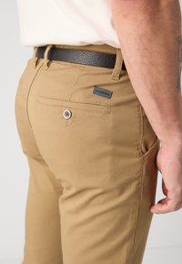 Khaki chinos med en jevn tekstur, med en baklomme med knappeåpning og et belte i læraktig svart materiale med strukturert overflate.