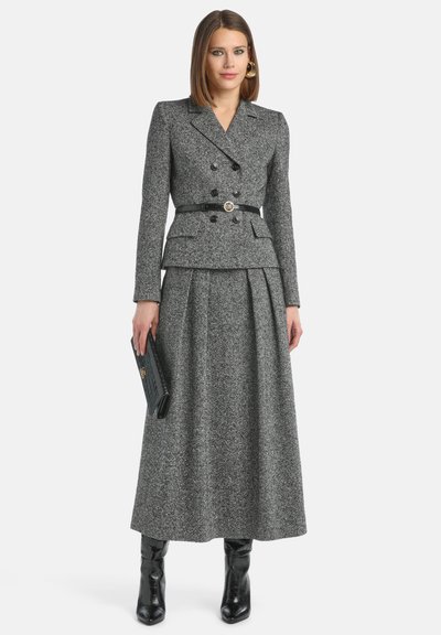 Costume en tweed gris avec une veste cintrée à double boutonnage, taille peplum et jupe longue. Ceinture noire avec boucle ronde et bottes à talons assorties.
