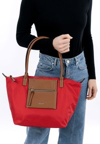 Red nylon tote bag with brown leather accents and a front zip pocket. Features dual handles, a smooth texture, and a spacious design.