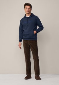 Marineblauwe hoodie met een voorzak en logo; gecombineerd met bruine corduroybroeken en bruine leren schoenen. Gladde stoftextuur.