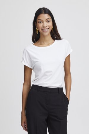 Woman with long dark hair wearing gold fan-shaped earrings, white short-sleeve shirt, and black trousers, smiling with one hand in pocket.