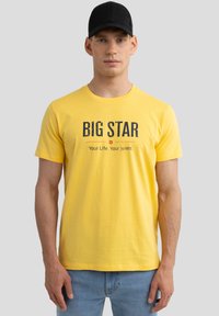 Dzeltena kokvilnas T-krekls ar "BIG STAR" tekstu tumši zilā krāsā, ar apaļu kakla izgriezumu. Modelis valkā gaiši zilo džinsus.